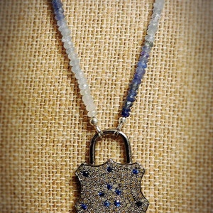 May include: A silver lock pendant necklace with a blue and white ombre gemstone bead chain. The lock is covered in small, clear gemstones and has a few blue gemstones in the center.