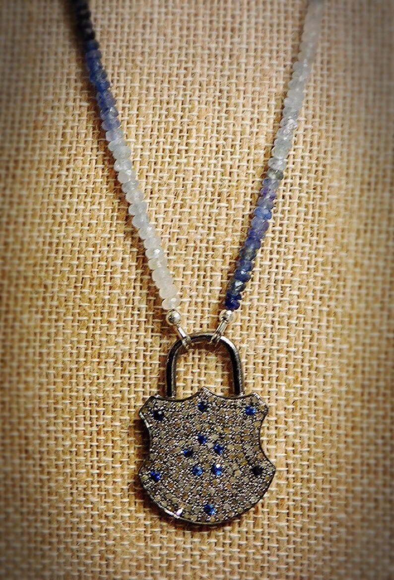 May include: A silver lock pendant necklace with a blue and white ombre gemstone bead chain. The lock is covered in small, clear gemstones and has a few blue gemstones in the center.