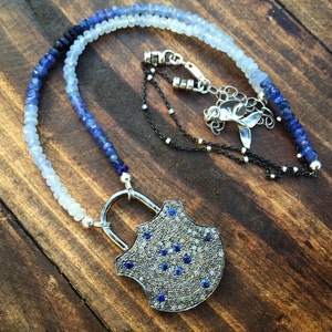 May include: A silver necklace with a padlock pendant set with blue sapphires and diamonds. The necklace is made of blue and white beads.