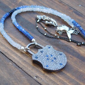 May include: A silver padlock pendant necklace with a blue and white gemstone bead chain. The padlock is encrusted with small diamonds and blue sapphires.