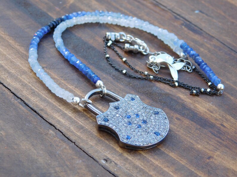 May include: A silver padlock pendant necklace with a blue and white gemstone bead chain. The padlock is encrusted with small diamonds and blue sapphires.