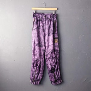 May include: Purple trousers with a tie-dye pattern and zipper accents. The trousers have a drawstring waist and are hanging on a wooden hanger.