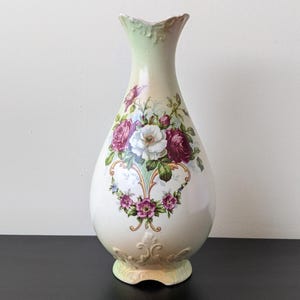 Könnte beinhalten: Eine dekorative Keramikvase mit cremefarbenem Körper und ausgestelltem Oberteil. Die Vase hat ein Blumendesign mit roten, lila und weißen Rosen. Der Sockel und die Oberseite haben geprägte Details, und die Oberseite ist hellgrün.