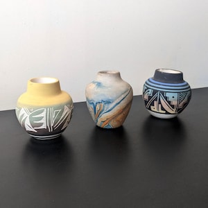 Peut inclure: Trois petits vases décoratifs en céramique. Le vase de gauche est jaune avec un motif géométrique blanc et vert. Le vase du milieu présente un motif marbré bleu et marron. Le vase de droite a un motif géométrique noir, bleu et blanc.