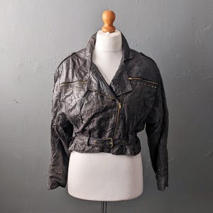 Könnte beinhalten: Eine dunkel anthrazitfarbene Lederjacke mit knittriger Textur, ausgestattet mit einem asymmetrischen goldfarbenen Reißverschluss, einem Taillengürtel mit Schnalle sowie kleinen goldfarbenen Metallnieten an den Brusttaschen und Ärmelbündchen.