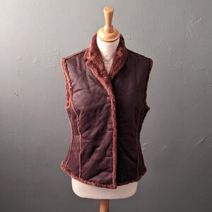 May include: A sleeveless waistcoat in a deep dark brown colour with a textured finish. The garment features a notched collar and visible seams along the front panels, all trimmed with a matching brown faux fur lining that is visible along the edges.