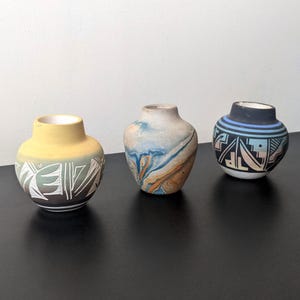 May include: Three small ceramic vases with decorative designs. The vases are painted in yellow, blue, and black, with geometric and abstract patterns. The vases are displayed on a black surface.