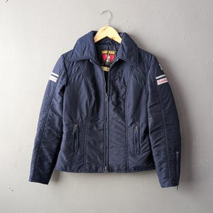 Könnte beinhalten: Eine marineblaue Reißverschlussjacke mit großem Kragen, zwei Reißverschlusstaschen vorne und weiß-roten Streifendetails an den Oberärmeln. Das Innenetikett zeigt den Text Innsbruck mit einem roten Adleremblem auf schwarzem Hintergrund.