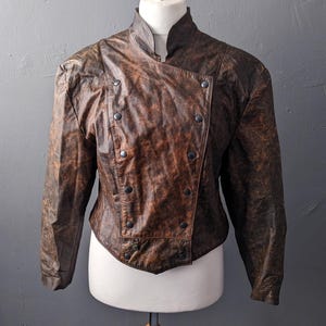 Puede incluir: Una chaqueta de cuero marrón con cierre de doble botonadura y cuello alto. La chaqueta tiene un aspecto desgastado y se muestra en un maniquí.