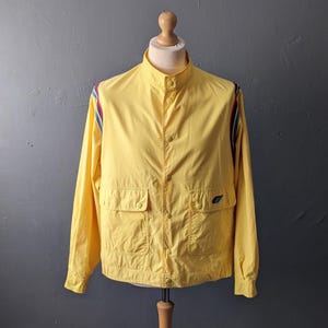 Può includere: Una giacca giallo brillante con colletto alla coreana, chiusura frontale con bottoni e due tasche con patta. Le spalle e le maniche sono decorate con strisce verticali rosse, bianche e blu. Un piccolo logo ricamato colorato si trova sulla tasca destra.