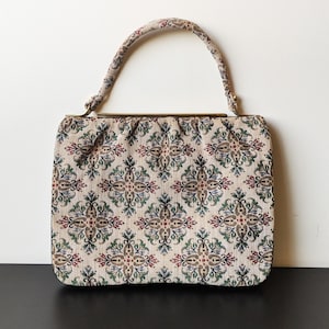 Peut inclure: Sac à main vintage beige avec un motif floral rouge, vert et bleu. Le sac a une forme structurée avec une poignée incurvée et un fermoir doré. Le tissu a une texture tissée et le motif est symétrique.