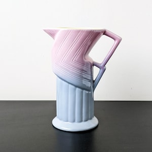 Può includere: Una brocca in ceramica caratterizzata da una sfumatura di colore che passa dal blu chiaro alla base al rosa tenue sulla parte superiore. La sezione inferiore presenta scanalature verticali, mentre il corpo superiore ha un motivo a chevron.