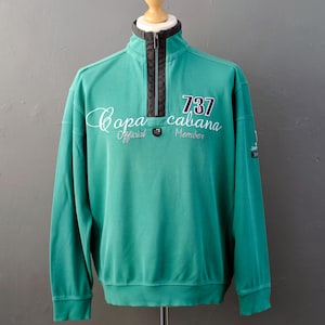 May include: A teal long-sleeve quarter-zip pullover with a textured ribbed fabric. The front features white embroidered text reading "Copa cabana Official Member" and "737" in a bold font. A dark grey trim accents the collar and zip area.