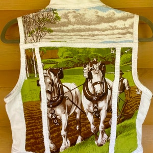Puede incluir: Chaleco vaquero blanco con un panel de tela vintage que presenta una escena pastoral de dos caballos arando un campo. El chaleco tiene un cinturón de cuero marrón con hebillas plateadas y un cuello blanco.
