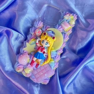 Peut inclure: Une coque de téléphone transparente décorée d'une texture de crème fouettée aux tons pastel, avec une figurine de Sailor Moon, un croissant de lune jaune, une tablette de chocolat rose, un petit beignet et une étoile. Le mot Sweet est inscrit.