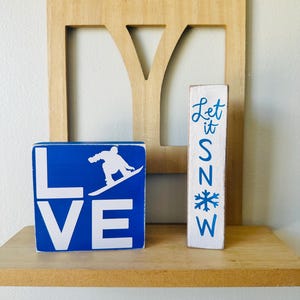 Peut inclure: Décor d'étagère en bois avec deux blocs peints et un dossier de chaise en bois. Un bloc bleu épelle "LOVE" avec un graphisme de snowboarder. L'autre bloc blanc dit "Let it Snow" en écriture bleue et un graphisme de flocon de neige.