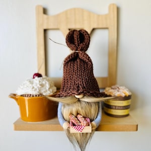 May include: A whimsical gnome figurine with a brown knitted hat and long, flowing beard, holding a miniature wooden tray with bread. The shelf also displays a cupcake with whipped cream and a pastry.