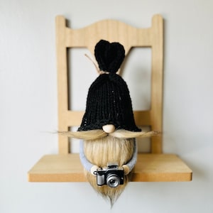 May include: A black knit hat with a long blonde beard and mustache gnome holding a small black camera. The gnome is sitting on a wooden shelf.