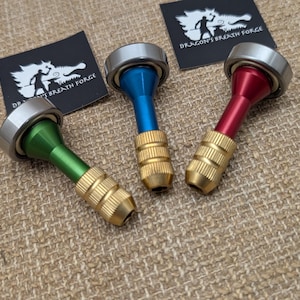 May include: Three metal spinning tops with ball bearings at the top, featuring green, blue, and red anodized aluminum stems with textured brass grips at the base. Two black square labels with the white text Dragon's Breath Forge are present.