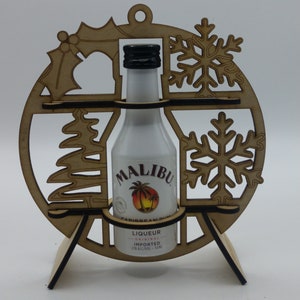 Könnte beinhalten: Weihnachtsbaumschmuck aus Holz in Form eines Kreises mit einer Flasche Malibu-Rum im Inneren. Der Schmuck ist mit einem Schneeflocken-, einem Weihnachtsbaum- und einem Stechpalmenmotiv verziert.