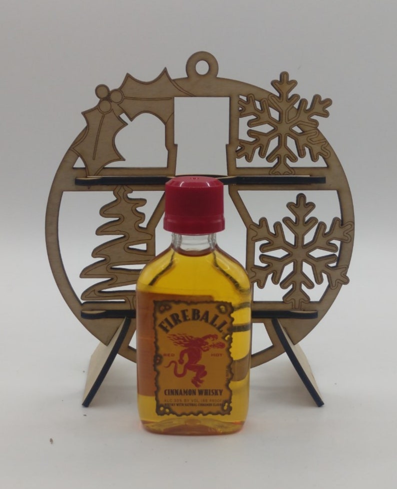 May include: A miniature bottle of Fireball Cinnamon Whisky in a wooden Christmas ornament shaped like a snowflake. The bottle is clear with a red cap and a red label with a red devil logo. The label reads "Fireball Cinnamon Whisky".