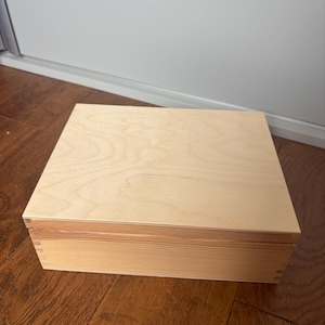 May include: A square wooden box with a light tan finish and visible wood grain patterns. The box features a flat lid and straight sides with visible finger joint construction at the corners, showcasing a natural, unpainted light brown wood texture.
