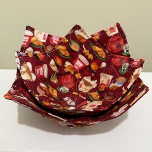 May include: Three nested, square-shaped fabric bowl cozies. The cozies are made of a burgundy fabric with a repeating pattern of Chinese food items, including takeout containers, dumplings, and tea cups. The edges are stitched.