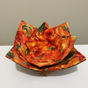 May include: A set of two bowl cozies with a peach pattern. The cozies are made of fabric with a repeating design of ripe peaches and green leaves. The bowl cozies are orange, red, and yellow.