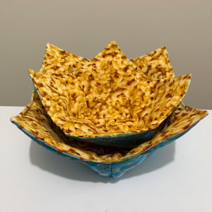 May include: Two fabric bowls, one nested inside the other. The bowls have a blue exterior with orange stitching and a pattern of macaroni and cheese on the interior. The bowls are made of fabric.