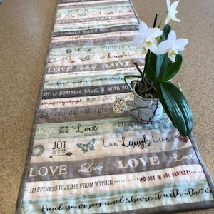 May include: A gray and white table runner with a striped pattern and the words "Live Laugh Love" and "Happiness Blooms From Within". A white orchid plant is sitting on top of the runner.