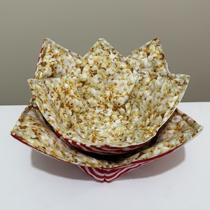 May include: Two fabric popcorn bowls. The interior fabric has a popcorn kernel pattern in shades of white, brown, and tan. The exterior fabric is red and white striped. The bowls are nested, with one inside the other.