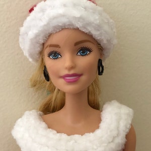 May include: A doll wearing a red and white knit hat with a white fur trim and a red and white knit dress with a white fur trim. The doll has blonde hair and blue eyes.