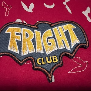 Op de afbeelding: Geborduurde patch met de woorden "FRIGHT CLUB" in geel en wit, omlijnd in zwart en bruin. De patch heeft de vorm van een vleermuis en staat op een rode achtergrond met witte vleermuis- en maanontwerpen.