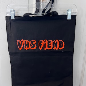 May include: Black tote bag with the words "VHS FIEND" in bright orange lettering. The bag has black handles and is hanging on a clear plastic hanger. The tote bag is made of a sturdy fabric.