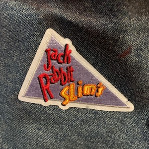May include: A triangular patch with the text "Jack Rabbit Slim's" in red and gold lettering. The patch has a white border and a purple background, and is sewn onto a denim jacket.