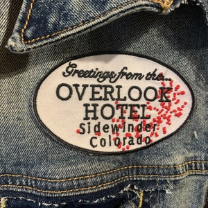 Peut inclure: Écusson brodé sur une veste en jean. Le patch ovale porte l'inscription "Greetings from the... OVERLOOK HOTEL Sidewinder Colorado" en noir, avec des détails brodés rouges. Le patch est cousu sur la veste.