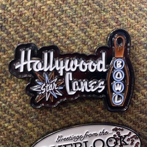 May include: Black and white enamel pin with the words "Hollywood Star Lanes" and a bowling pin design. The pin has a starburst graphic and is set against a textured background.