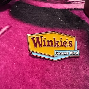 Könnte beinhalten: Rechteckiger Emaille-Pin mit dem Text "Winkie's" in kastanienbraunen Buchstaben auf gelbem Hintergrund. Darunter befindet sich der Text "Sunset Blvd" auf hellblauem Hintergrund. Der Pin hat einen goldenen Rand.