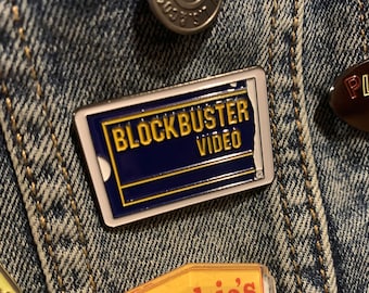 May include: A rectangular enamel pin featuring the Blockbuster Video logo in navy blue and gold on a white background. The pin is set against a denim jacket.