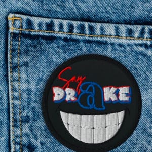 Peut inclure: Un patch smiley noir et blanc avec le texte "Say Drake" en lettres rouges, bleues et blanches cousu sur une poche en jean bleu.