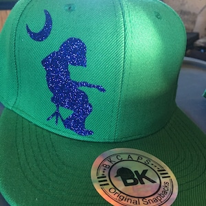 May include: Green snapback hat with a blue glitter design of a person playing a stringed instrument and a crescent moon. The hat has a flat brim and a round sticker that says "BK CAPS Original Snapbacks."