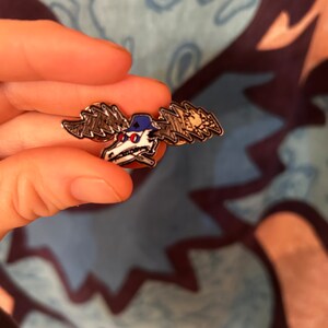 Puede incluir: Un pin de esmalte plateado con un alce de dibujos animados que lleva una gorra de b&eacute;isbol azul con un dise&ntilde;o de rayo blanco. El alce tiene ojos rojos y una calavera blanca con un dise&ntilde;o de huesos cruzados negros.