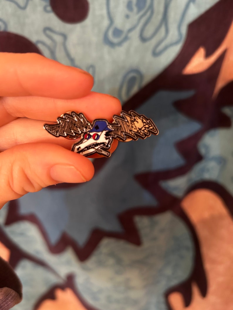 Puede incluir: Un pin de esmalte plateado con un alce de dibujos animados que lleva una gorra de b&eacute;isbol azul con un dise&ntilde;o de rayo blanco. El alce tiene ojos rojos y una calavera blanca con un dise&ntilde;o de huesos cruzados negros.