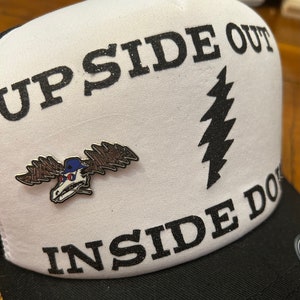 Puede incluir: Una gorra de camionero blanca con letras negras de purpurina que dicen "UP SIDE OUT" e "INSIDE DOWN". Un gr&aacute;fico de rayo negro est&aacute; en la parte delantera de la gorra. Un pin de alce plateado est&aacute; sujeto a la gorra.