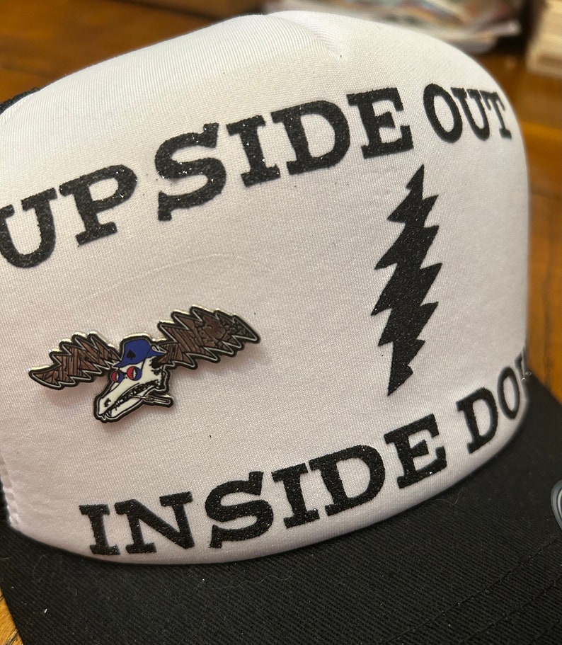 Puede incluir: Una gorra de camionero blanca con letras negras de purpurina que dicen "UP SIDE OUT" e "INSIDE DOWN". Un gr&aacute;fico de rayo negro est&aacute; en la parte delantera de la gorra. Un pin de alce plateado est&aacute; sujeto a la gorra.