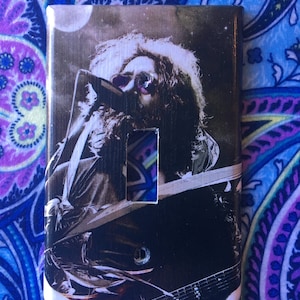 May include: A light switch cover featuring a black and white photo of a musician playing a guitar. The musician has long hair and a beard. The background is a blurry image of a stage.