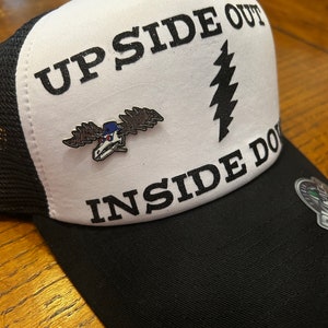 Puede incluir: Una gorra de camionero blanca y negra con el texto "UPSIDE OUT INSIDE DOWN" y un gr&aacute;fico de rayo negro y blanco. Un peque&ntilde;o pin de metal con un alce de dibujos animados que lleva un sombrero est&aacute; sujeto a la parte delantera de la gorra.