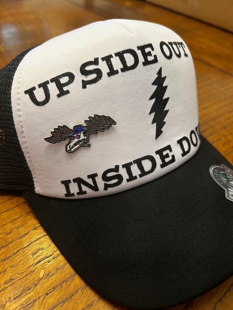 Puede incluir: Una gorra de camionero blanca y negra con el texto "UPSIDE OUT INSIDE DOWN" y un gr&aacute;fico de rayo negro y blanco. Un peque&ntilde;o pin de metal con un alce de dibujos animados que lleva un sombrero est&aacute; sujeto a la parte delantera de la gorra.