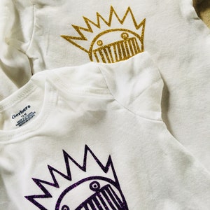 May include: Two white baby onesies with a graphic of a cartoon creature with a crown. The onesie on top has a gold glitter creature and the onesie on the bottom has a purple glitter creature. The onesies have the Gerber brand label.