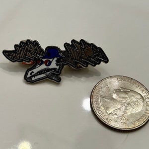 Puede incluir: Un pin de esmalte plateado con un cr&aacute;neo de dibujos animados que lleva una gorra de b&eacute;isbol azul y gafas de sol. El cr&aacute;neo tiene un dise&ntilde;o de rayo en cada lado de su cabeza. El pin es aproximadamente del tama&ntilde;o de una moneda de 20 c&eacute;ntimos de euro.